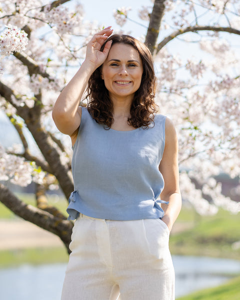 A smiling woman with dark, wavy hair wears a cloudy blue sleeveless linen top with side ties, paired with white linen pants. She stands outdoors under a blooming cherry blossom tree, with a body of water and green landscape in the soft background.