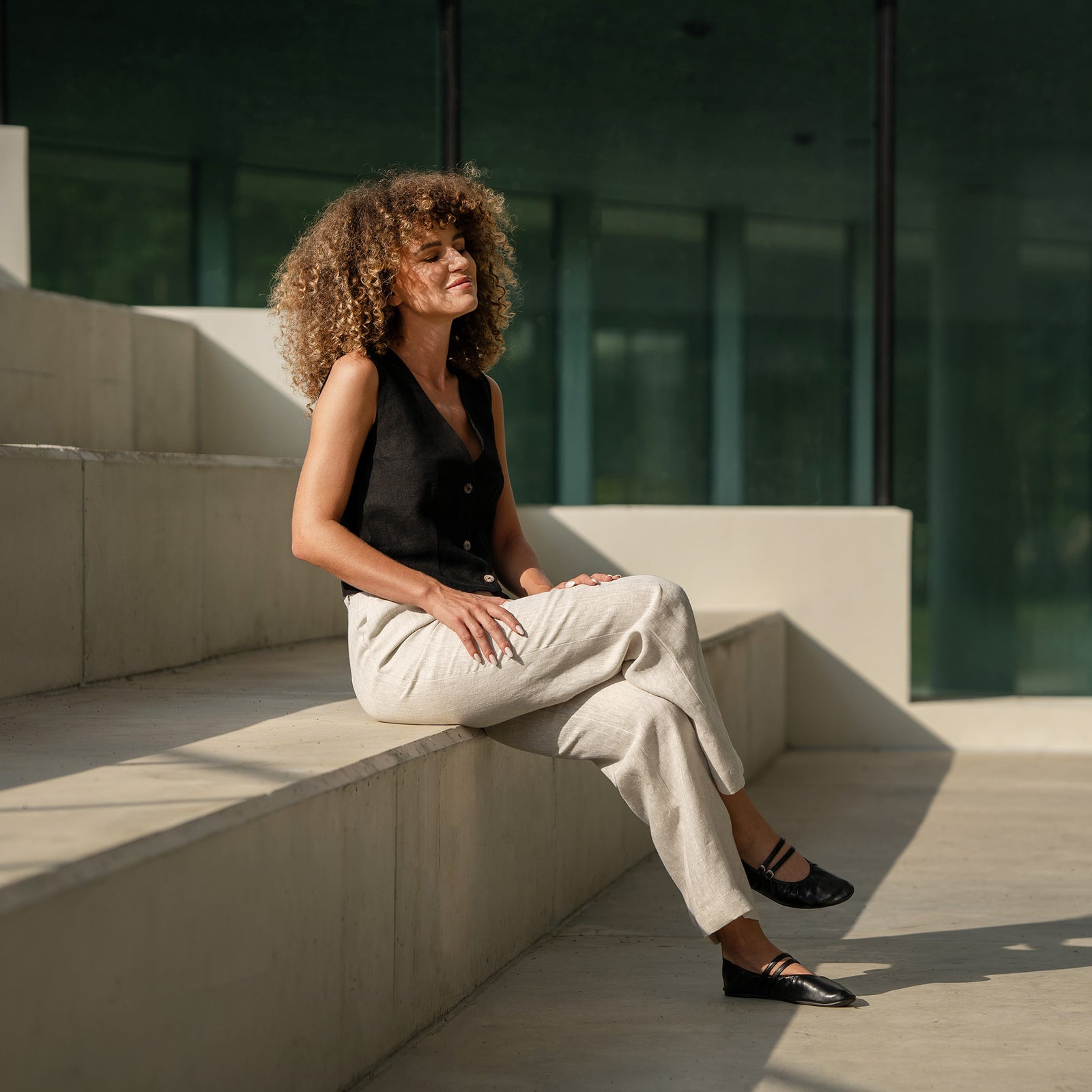 A person is shown from the waist down, modeling stone green, high-waisted, linen wide-leg trousers. They are wearing black open-toe sandals with thin straps and have their hands on their hips. The setting appears to be outdoors on an asphalt path, with a black metal fence and greenery visible in the background.