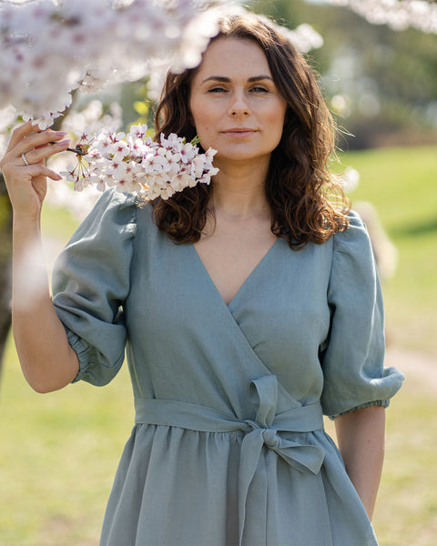 A woman with brown hair, wearing a mint green linen wrap dress with elbow-length puff sleeves, stands outdoors next to a tree with white blossoms.