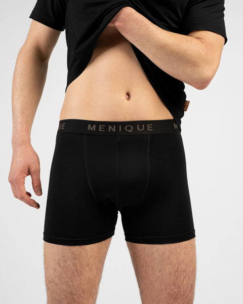 A man wearing a black boxer briefs. He is holding the bottom edge of his t-shirt with both hands, revealing the branded waistband of the underwear.