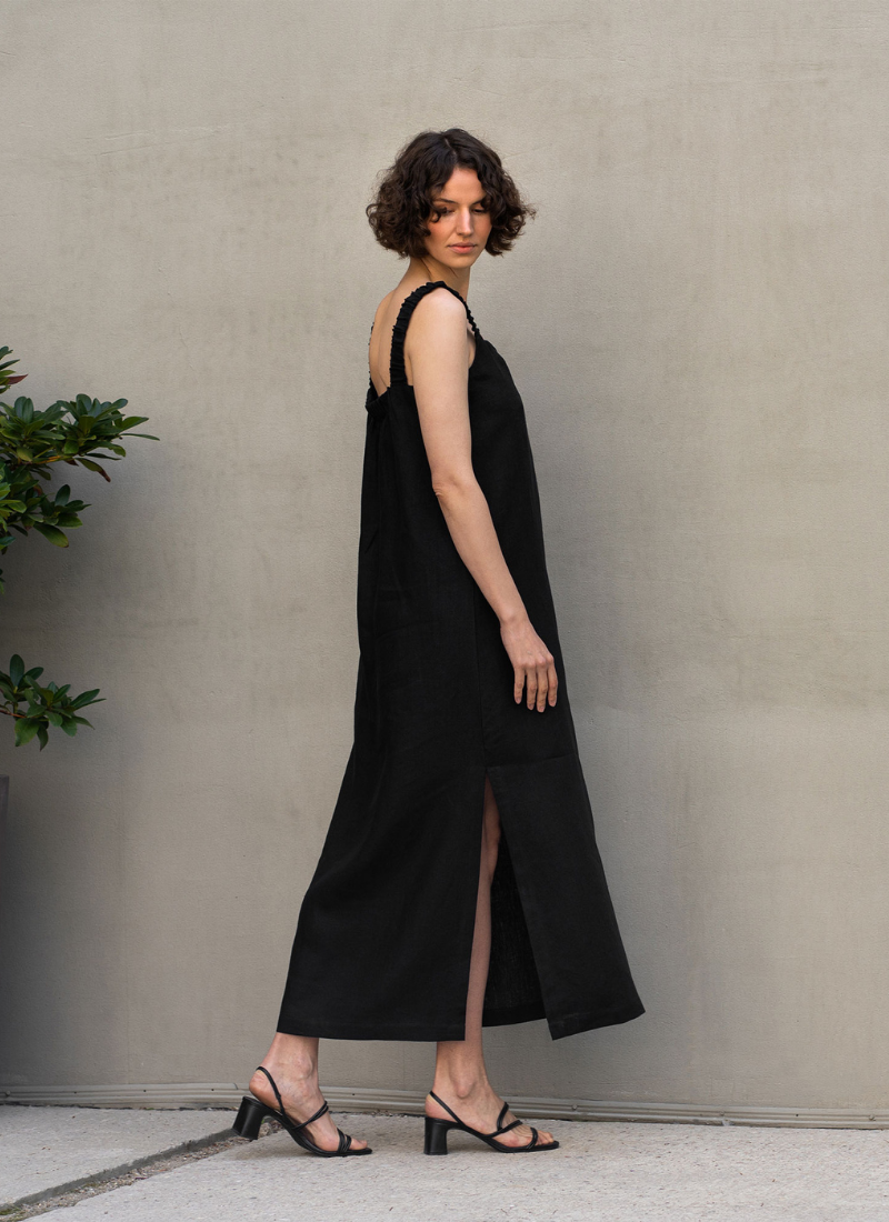 Woman wearing a minimalist black sleeveless midi dress with a side slit, standing against a neutral wall, styled with black heeled sandals for an elegant, modern look.