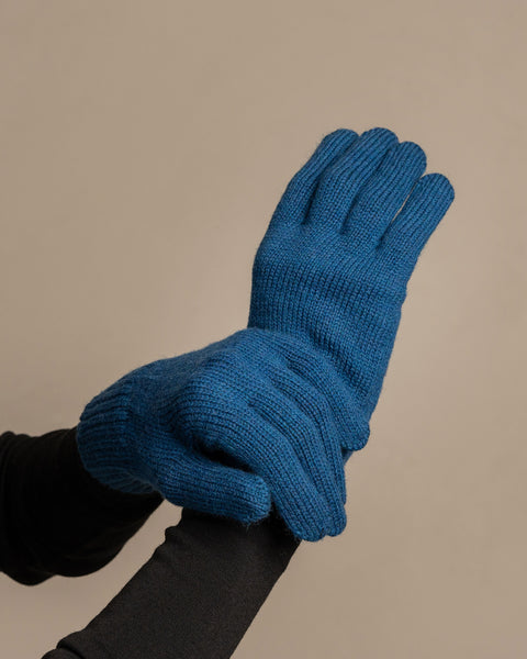 A close-up shot of a person&#39;s hands wearing menique brand vibrant blue, ribbed knit gloves. The right hand is gently supporting the underside of the left hand, which is slightly cupped. The person is wearing a dark long-sleeved shirt underneath. The background is a solid, neutral gray.