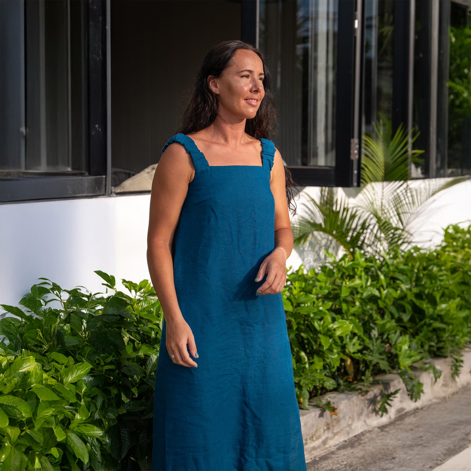 A woman with long dark hair, wearing a sleeveless, linen cobalt blue maxi dress with a square neckline and tie straps at the shoulders, stands barefoot on a grassy cliff overlooking a rocky coastline and ocean. She has one hand raised near her face and is looking out towards the water. The background features lush green vegetation and some buildings on the cliffs. The sky is overcast.
