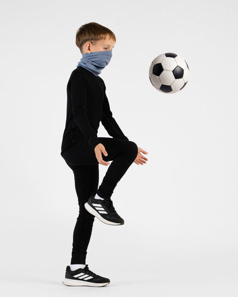A young boy stands in profile facing right against a white background, demonstrating a soccer move by juggling a black and white soccer ball with his knee. He is wearing a black base layer set, black and white Adidas sneakers, and a light blue neck gaiter pulled over his nose and mouth.