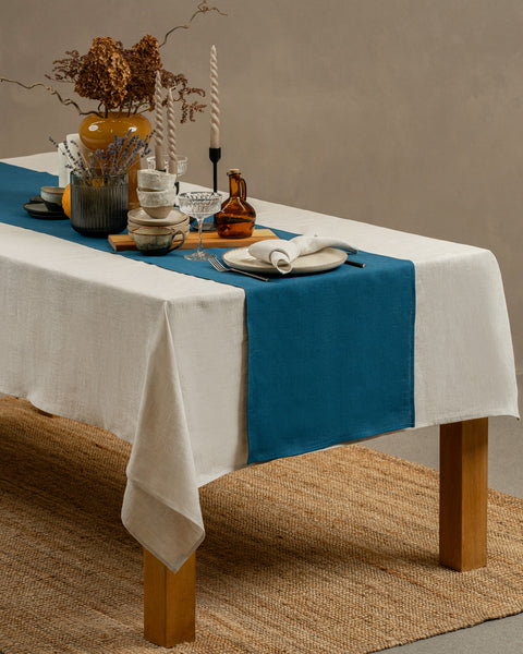 A view of a wooden table covered with a white tablecloth, layered with a cobalt blue table runner on top. There are vases, mugs and other ceramics on top of it. The table is positioned on top of a carpet, in a studio background.