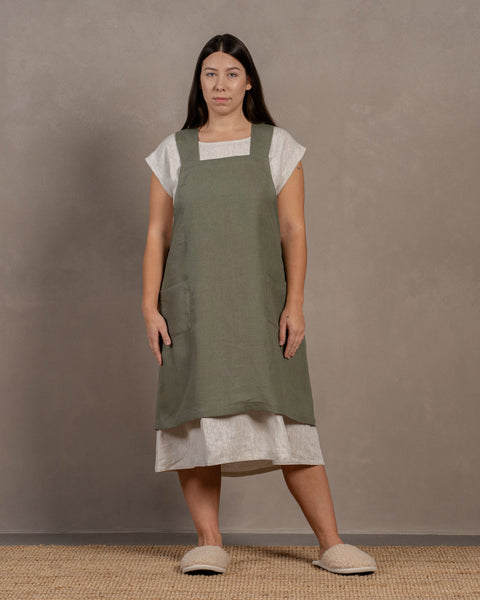 A woman with long dark hair stands facing the camera, wearing a menique stone green apron over a white linen dress. She also wears light-colored slippers and stands on a woven rug against a mottled gray background.