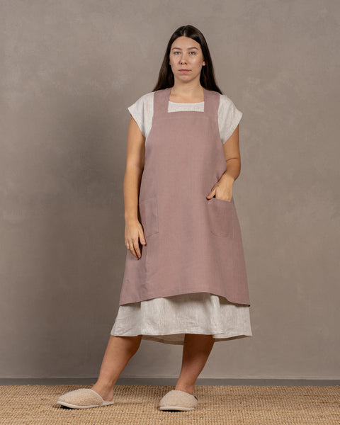 A young woman with dark hair standing in a studio background, with one hand inside the front pocket. She is wearing a linen dress with a linen cross-back japanese apron in faded rose color in size L/XL on top.