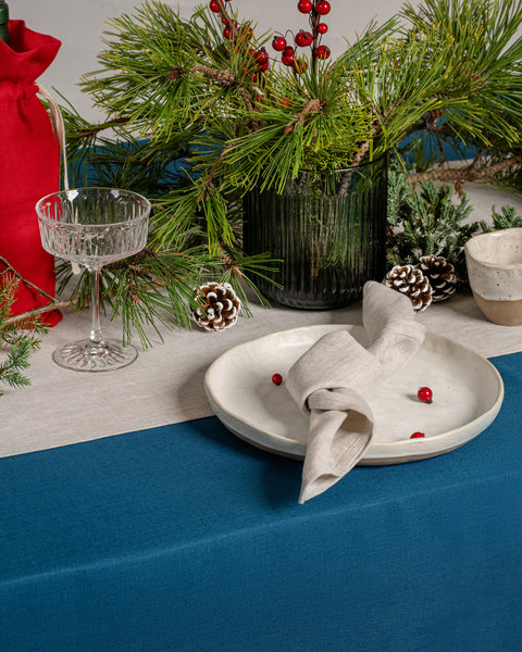 Christmas table setting with teal linen table runner, neutral linen napkin, red wine bag, pine greenery, and festive holiday décor.