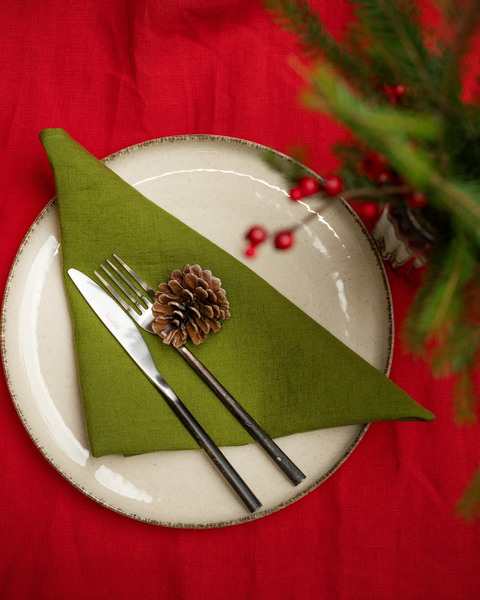 A festive table setting shown from above, featuring a cream-colored plate on a red tablecloth. A green linen napkin is folded into a triangle and topped with a pinecone. A fork and knife rest on the napkin, and evergreen branches with red berries frame the upper corner.