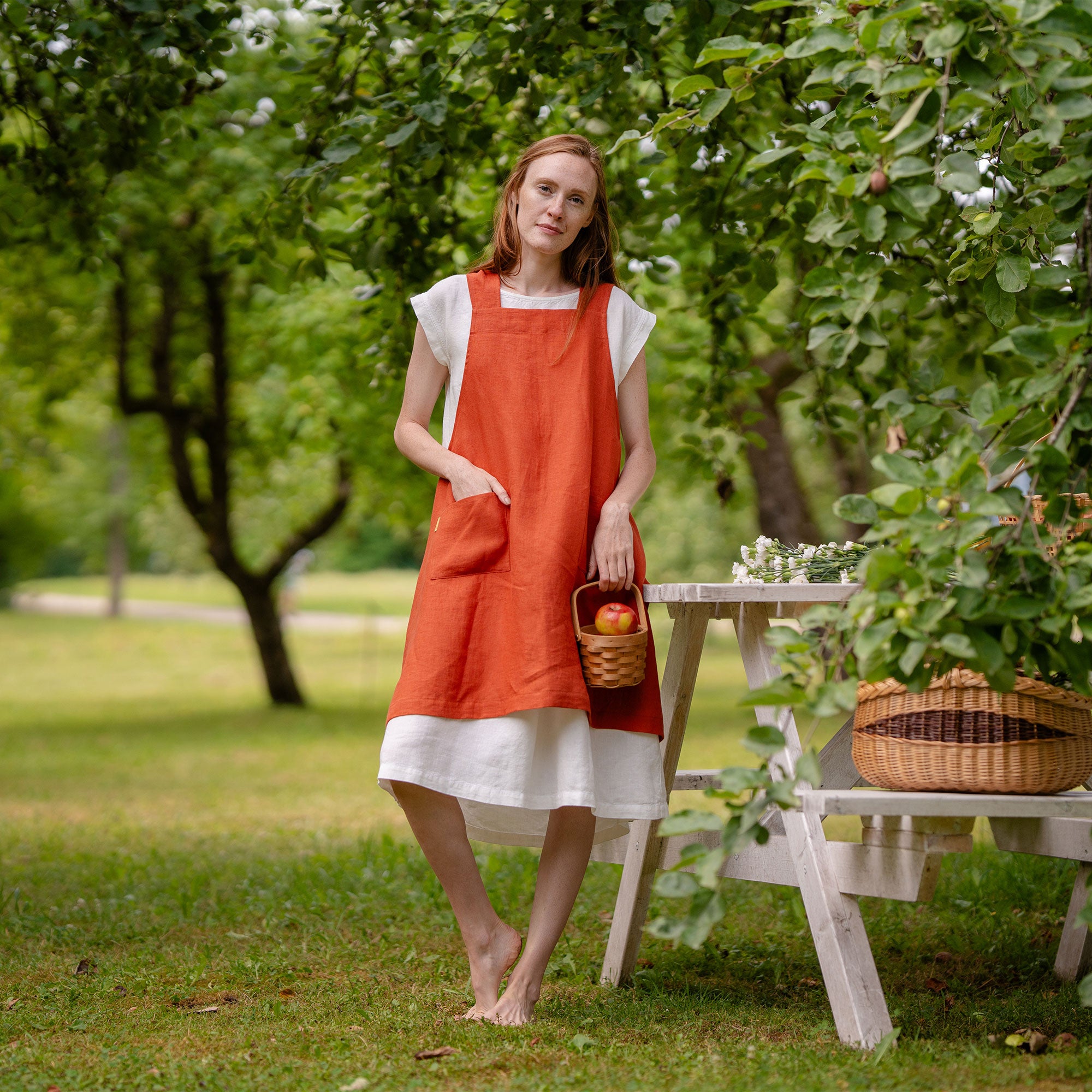 A woman standing and smiling in a warm, rustic kitchen while wearing a red Japanese-style apron. A wooden island with chopped greens and vegetables sits in front of her.