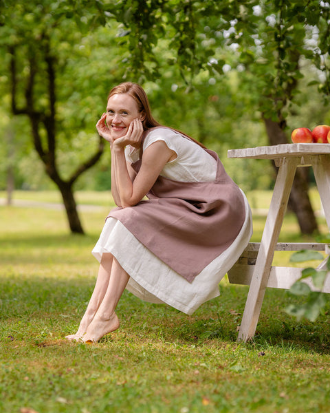 A fair-skinned woman with long reddish hair wearing linen menique apron sits barefoot on a white bench in a sunny garden, smiling with her chin resting on her hands. A small table with apples and flowers is beside them.
