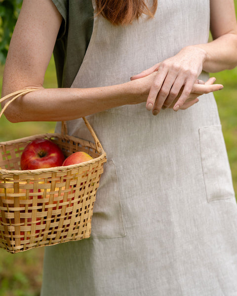 A close-up shot of a womans hands and torso, wearing a natural color menique linen apron, holding a woven basket containing red apples.