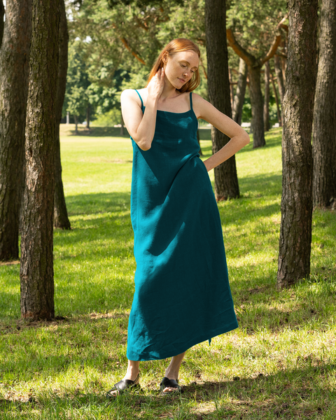 A fair-skinned woman with long, flowing reddish-brown hair stands in a sun-dappled forest clearing, wearing a sleeveless, floor-length menique linen dress in the color cobalt blue and black shoes. She is one hand gently touching her neck and the other resting on her hip, and her eyes are closed as she appear to be enjoying the peaceful natural setting. Tall trees with textured bark surround her, and sunlight filters through the leaves, casting soft shadows on the grassy ground.