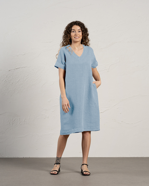 A woman standing against a plain background, wearing a loose, mid-length cloudy blue dress with a V-neckline and short sleeves. She has curly hair and is posing with a hand in her pocket.
