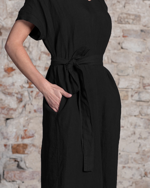 A woman models menique linen loose pure black dress with a tied waist and a hand in the side pocket, standing against a textured brick wall.