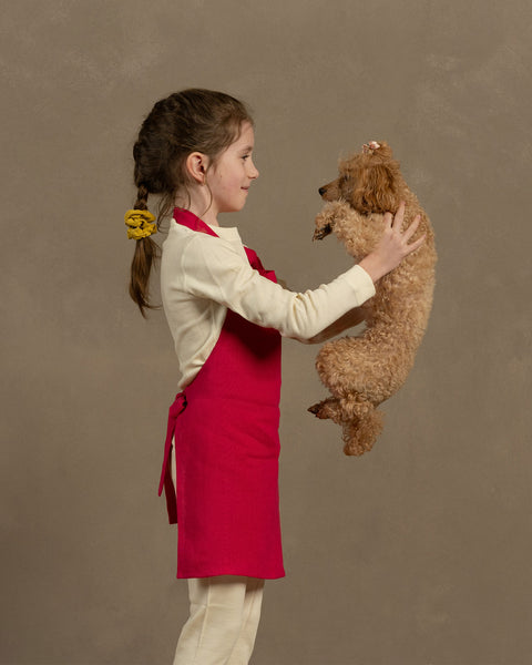 Young girl holding a small brown dog against a plain background wearing hot pink menique linen kids apron
