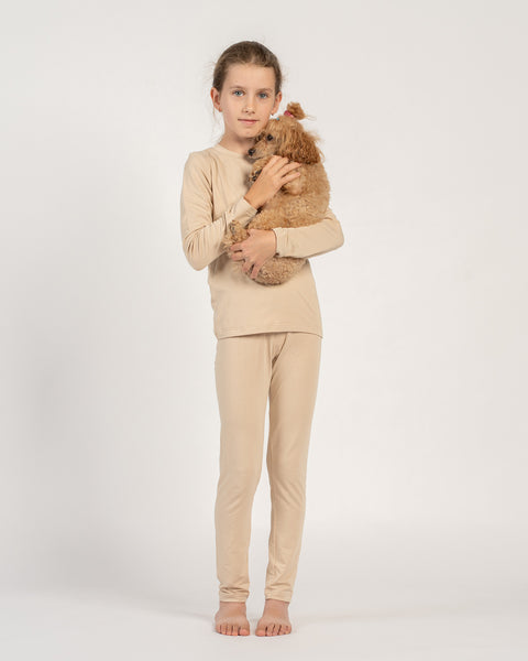 Girl wearing a beige bamboo long-sleeve top and bottoms, standing barefoot and holding a small brown poodle in her arms.