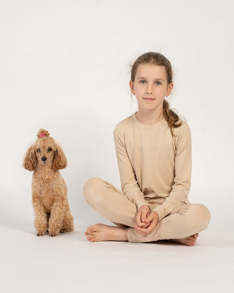 Girl sitting cross-legged in beige bamboo long-sleeve top and bottoms, next to a small brown poodle with a pink hair tie.