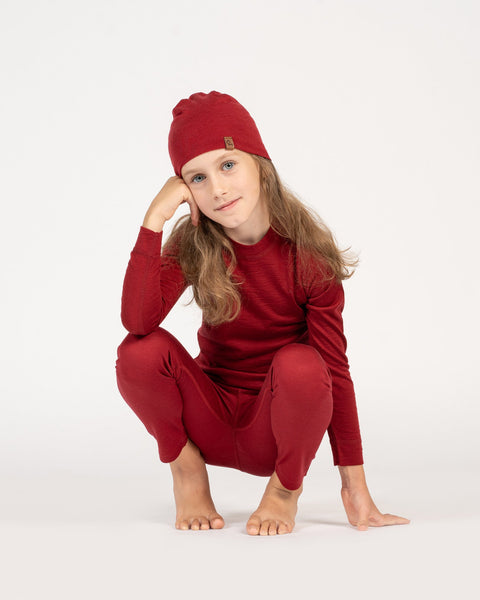 a girl, crouching down, wearing the kids&#39; merino beanie in the color royal cherry, and a matching color merino set with long sleeve top and pants.