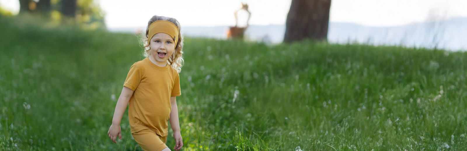 A young girl with curly blonde hair playfully sticking her tongue out, wearing a matching mustard yellow headband, short-sleeve t-shirt, and shorts while walking in a lush, green field.