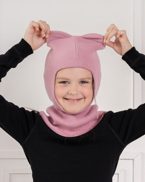 A smiling young girl wearing a black long-sleeved top and a pink knitted balaclava. She has her arms raised, playfully holding two small, cute animal-like "ears" knitted into the top of the balaclava. The background is a plain white wall with subtle paneling.