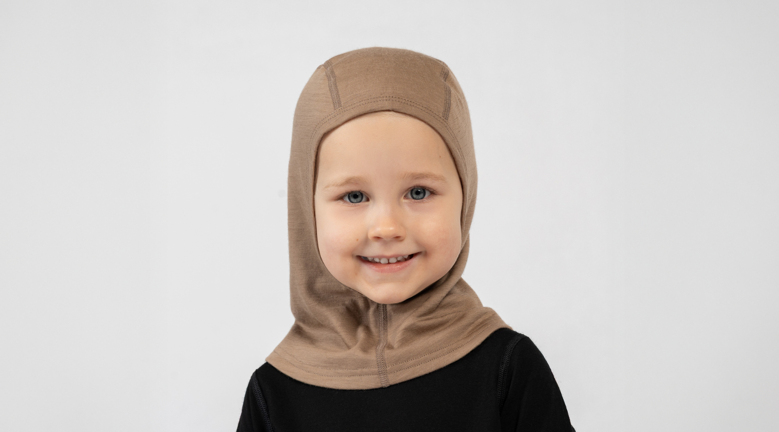 A young, smiling child standing against a white background. They are wearing a black long-sleeved top and a snug, beige balaclava that covers their head and neck while leaving their face exposed.