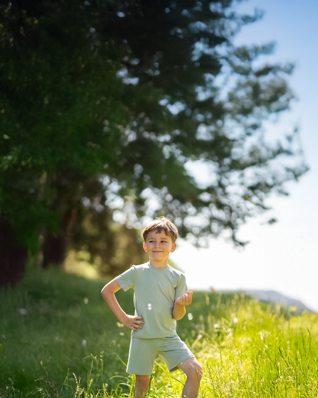 A young boy in a light green short-sleeve t-shirt and matching shorts standing confidently with his hands on his hips in a sunlit, grassy meadow.