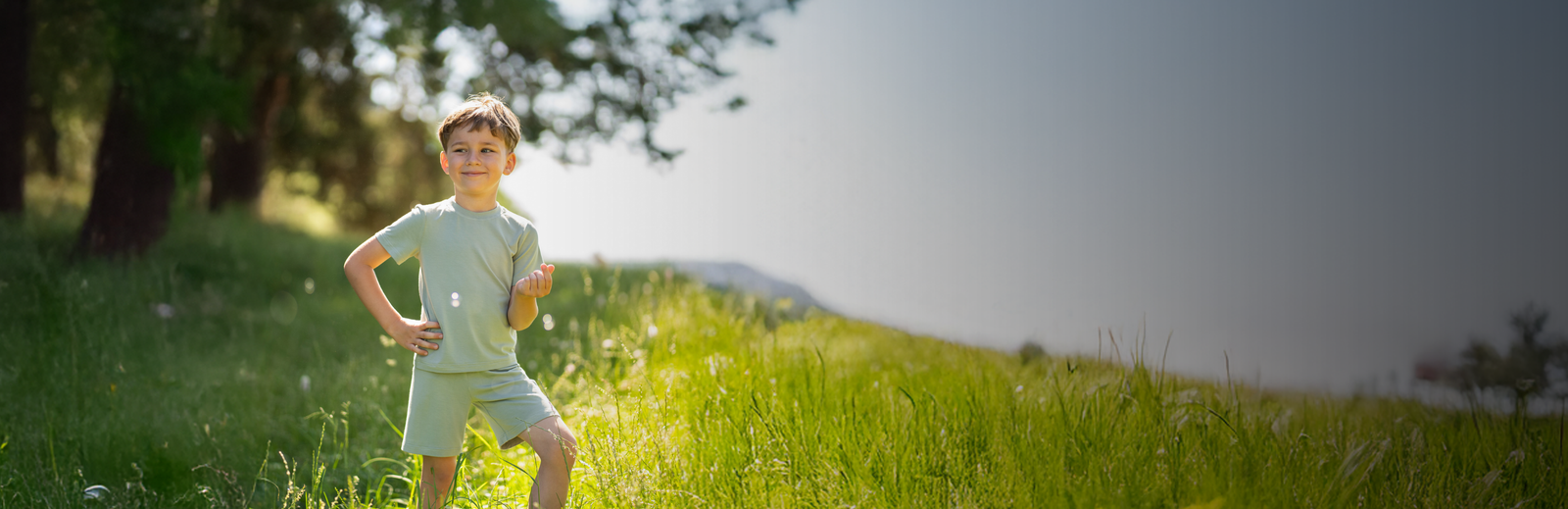 A young boy in a light green short-sleeve t-shirt and matching shorts standing confidently with his hands on his hips in a sunlit, grassy meadow.