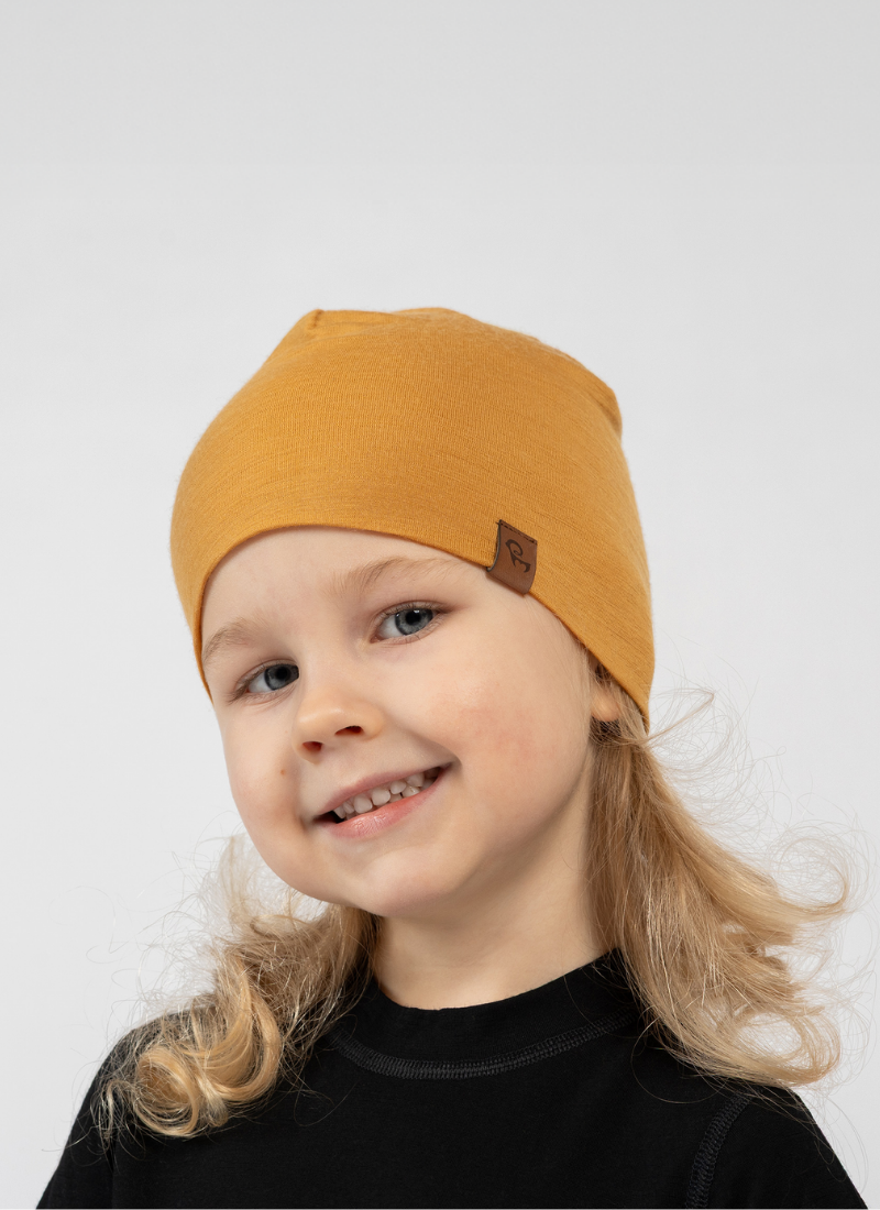 A young child with blonde curly hair smiling brightly at the camera against a white background. They are wearing a black top and a mustard yellow beanie featuring a small brown logo tag on the edge.