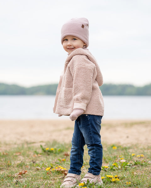 A young child is standing on a grassy and sandy shore next to a body of water, possibly a lake or large pond. The child is looking over their left shoulder and smiling at the camera. They are dressed in a light pink knit beanie with a small brown patch, a bulky light pink or beige sherpa-style jacket, dark blue jeans, and light-colored shoes. Some yellow wildflowers are scattered in the grass. The sky appears overcast.