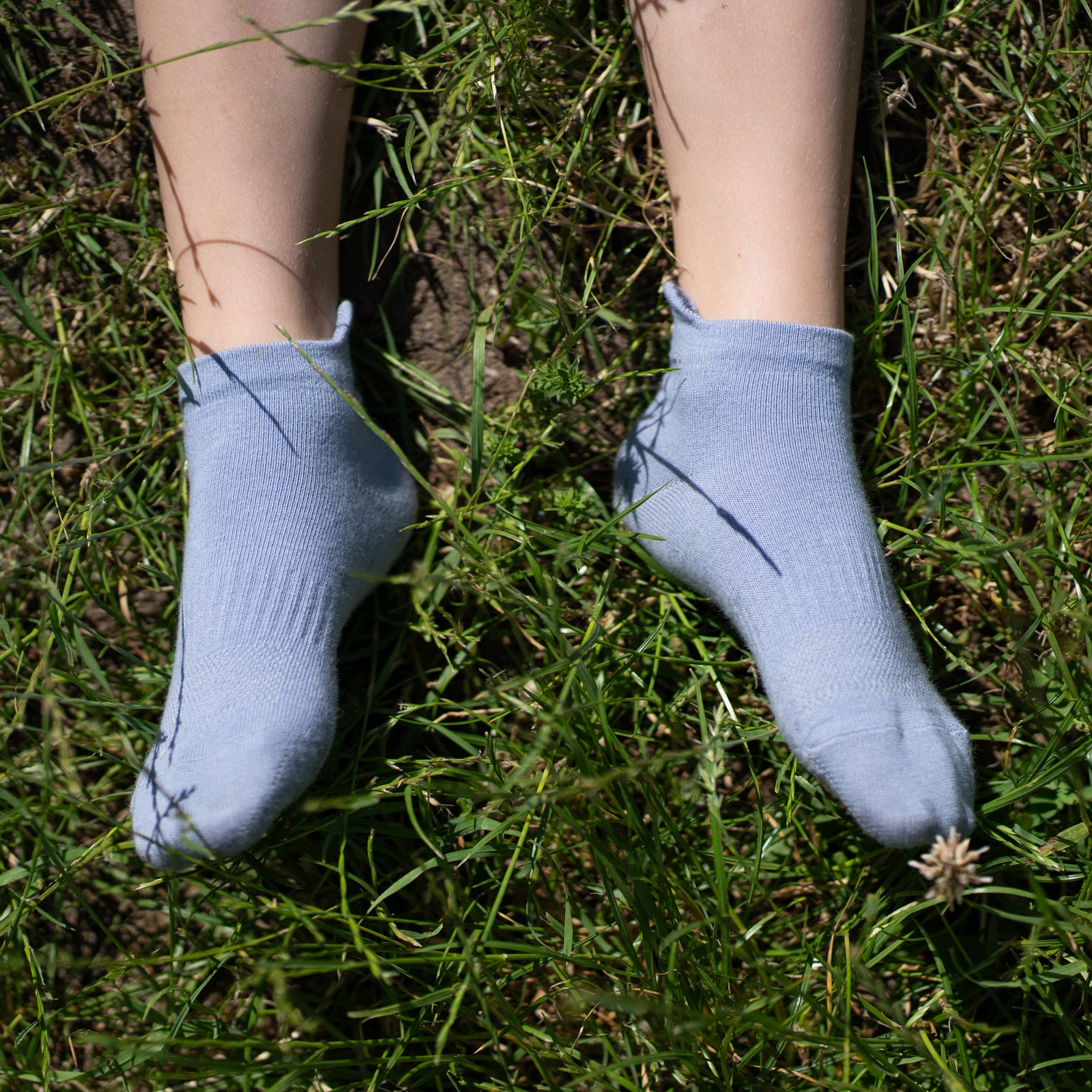 A close-up of two children's legs playfully resting together on a wooden park bench. One child is wearing light blue ankle socks, while the other wears dusty pink ankle socks with the word "MENIQUE" printed on them.