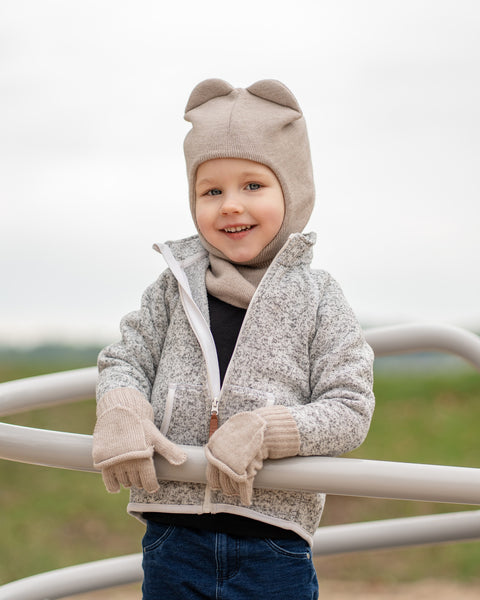 Child wearing a creamy beige knitted balaclava with bear-ear design and matching convertible gloves, smiling outdoors in a light gray jacket.