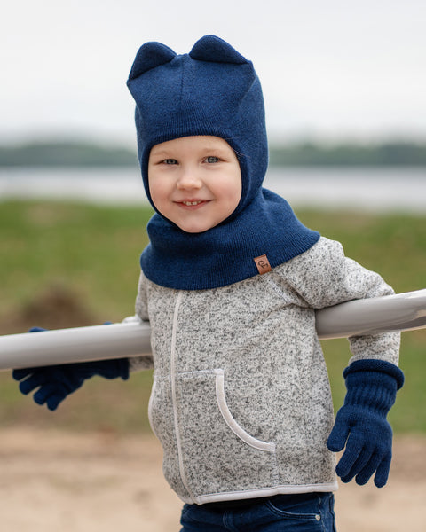 A young child with fair skin and light-colored hair is smiling at the camera, leaning on a light-colored horizontal bar outdoors. They are wearing a grey speckled zip-up jacket, blue jeans, and dark blue knitted menique accessories, including a balaclava-style hat with two rounded ears on top and a pair of ribbed gloves. In the soft-focus background, there&#39;s a body of water and a faint green shoreline.