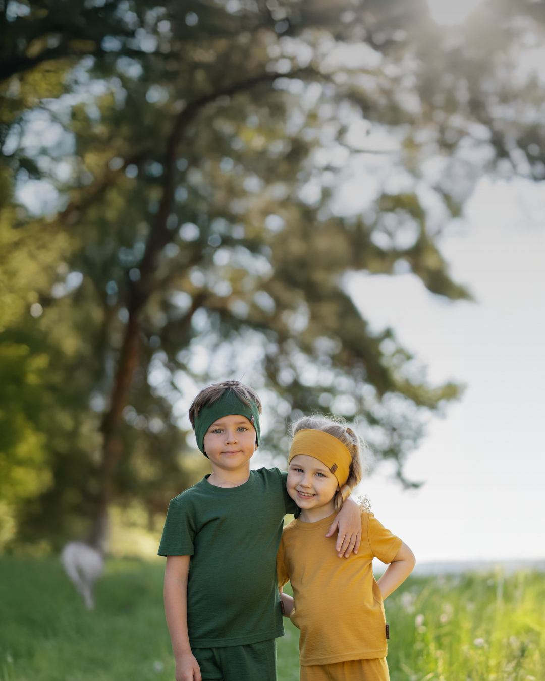 A young boy in a dark green outfit and matching headband standing with his arm warmly around a young girl in a mustard yellow outfit and headband in a bright, grassy field.