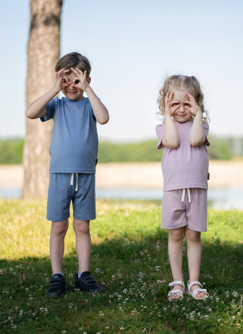 Two young children standing on a grassy field, playfully making binocular shapes over their eyes with their hands. The boy wears a dusty blue short-sleeve top and shorts set, and the girl wears a dusty pink short-sleeve top and shorts set.