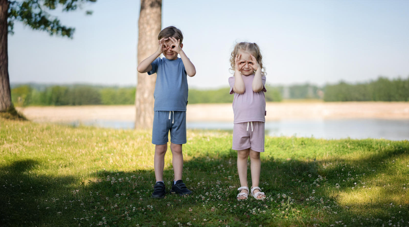 Two young children standing on a grassy field, playfully making binocular shapes over their eyes with their hands. The boy wears a dusty blue short-sleeve top and shorts set, and the girl wears a dusty pink short-sleeve top and shorts set.