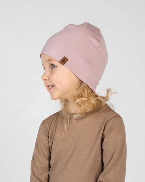 A studio portrait of a smiling young blonde girl looking off to her left in profile against a plain light grey background. She is wearing a brown long-sleeved top and a dusty pink beanie featuring a small brown logo tag.