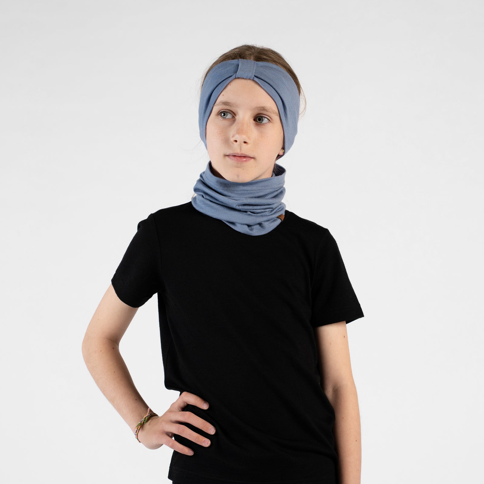 A close-up studio portrait of a young girl smiling directly at the camera. She wears a short-sleeved dusty blue top and a matching dusty blue ribbon headband. Her hands are gently resting behind her neck against a plain light grey background.