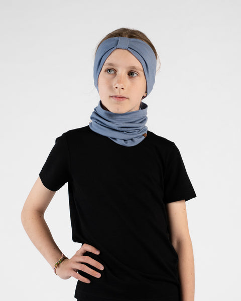 A studio portrait of a young girl standing with her hand on her hip, looking off to her left against a white background. She is wearing a black short-sleeved t-shirt, a dusty blue ribbon headband, and a matching dusty blue neck gaiter bunched loosely around her neck.