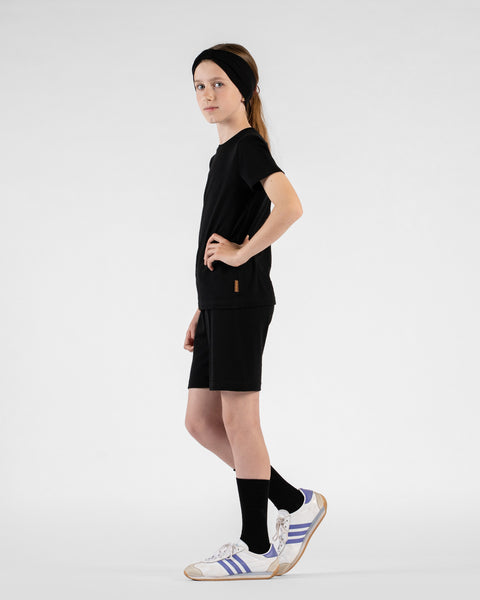 An older child poses in profile against a light background wearing a black short-sleeve t-shirt, black shorts, and a wide black headband. They have one hand on their hip and are looking back over their shoulder at the camera, completing the outfit with tall black socks and white striped sneakers.