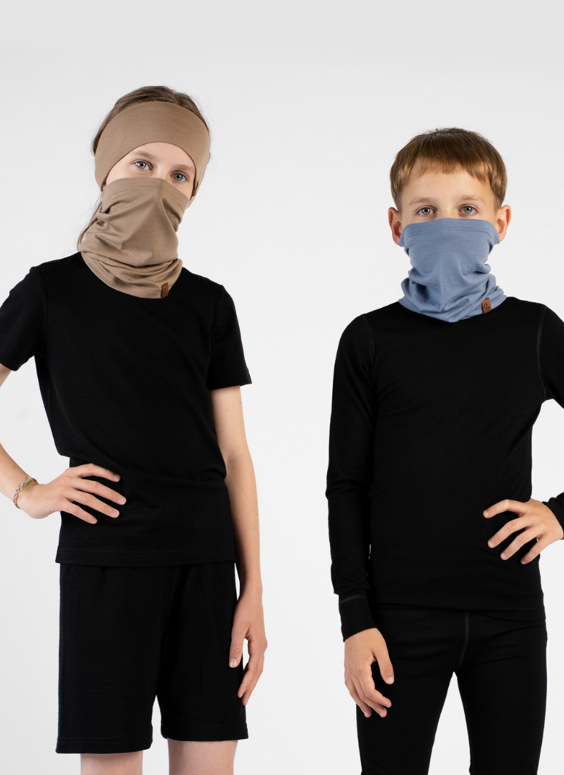 Two children standing side-by-side against a white background, wearing black short-sleeved tops. The girl on the left wears a beige headband and a matching beige neck gaiter pulled up over her nose and mouth. The boy on the right wears a dusty blue neck gaiter, also pulled up over his nose and mouth.
