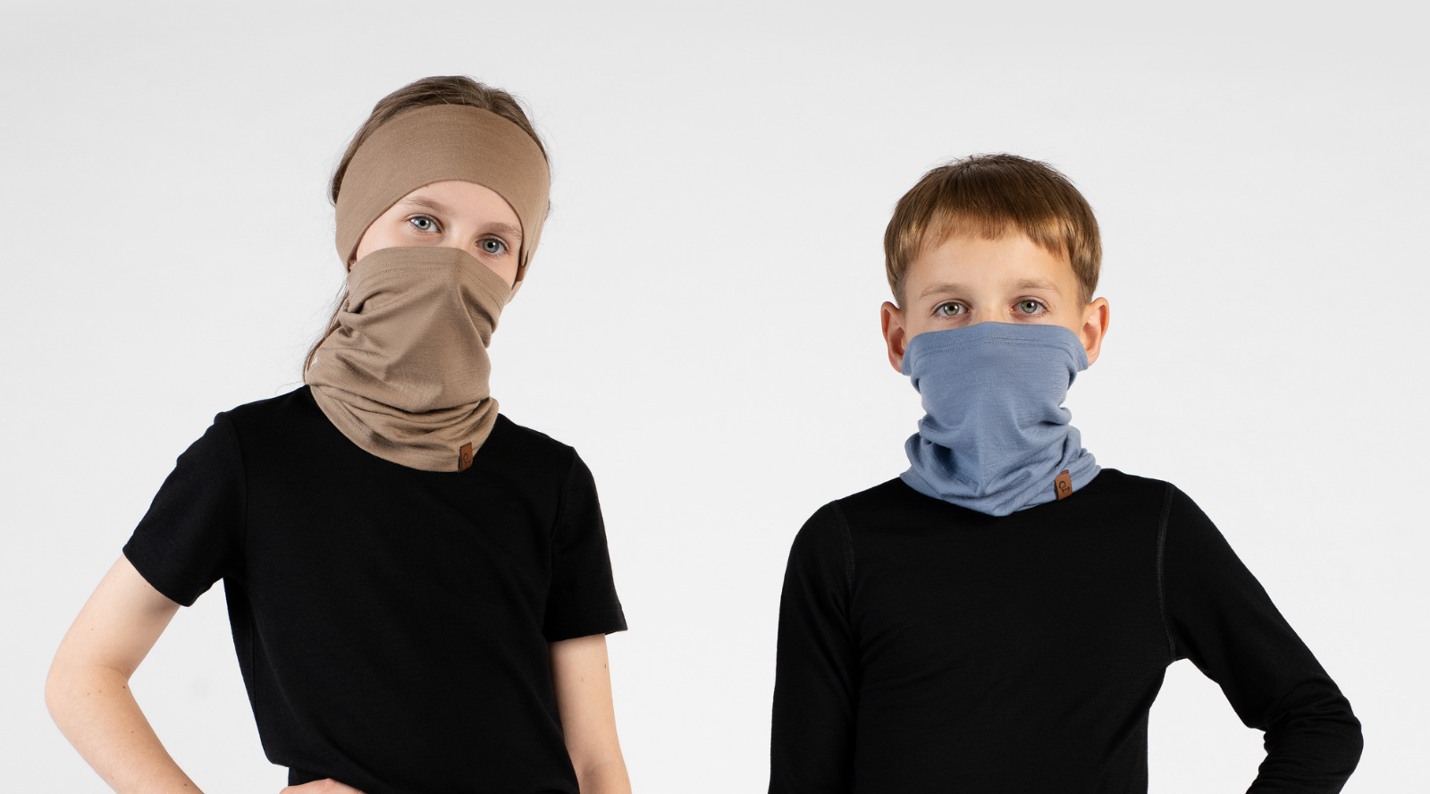 Two children standing side-by-side against a white background, wearing black short-sleeved tops. The girl on the left wears a beige headband and a matching beige neck gaiter pulled up over her nose and mouth. The boy on the right wears a dusty blue neck gaiter, also pulled up over his nose and mouth.
