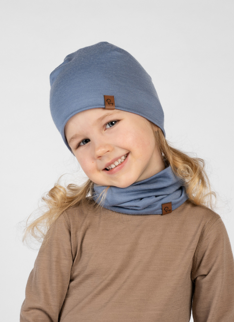 A young girl with blonde curly hair tilting her head and smiling at the camera against a white background. She is wearing a brown long-sleeved top, a dusty blue beanie, and a matching blue neck gaiter scrunched around her neck.