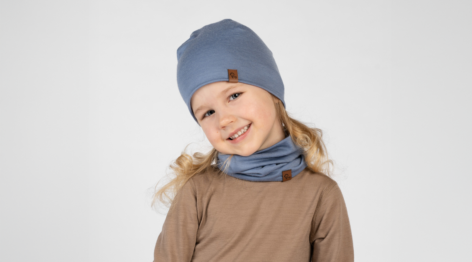 A young girl with blonde curly hair tilting her head and smiling at the camera against a white background. She is wearing a brown long-sleeved top, a dusty blue beanie, and a matching blue neck gaiter scrunched around her neck.