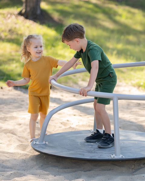 Two children playing together on a playground merry-go-round while wearing matching Merino wool T-shirts and shorts in yellow and dark green. The lightweight, breathable kids' outfits are showcased in a sunny outdoor setting, perfect for active play, everyday comfort, and family adventures.