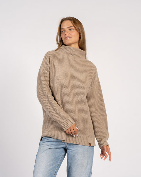 A woman posing in a beige knit sweater with a relaxed fit and long sleeves, slightly turning to the side.