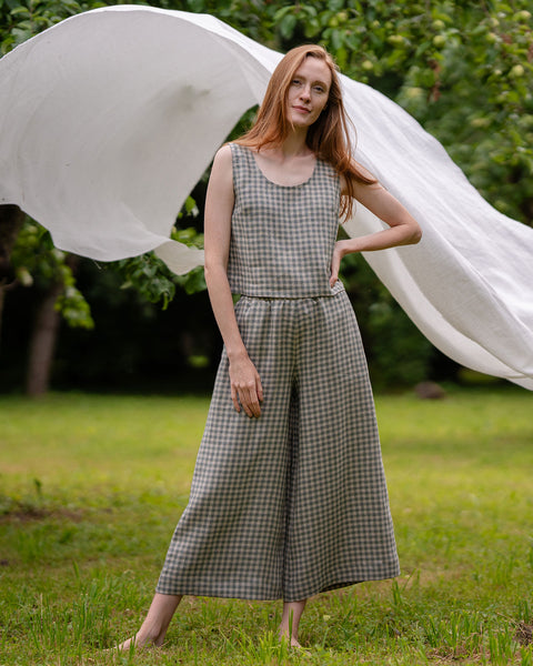 The model is posing in the green gingham LANA culottes with a matching ANA crop top. She is facing the camera, has her one hand on the hip, while the other one hangs by her side. She is relaxed, behind her is a white garment and nature views.