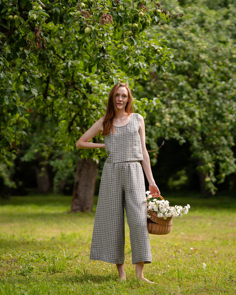 The model is facing the camera, while wearing the green gingham sleeveless blouse and culottes matching set. She is holding a basket full of wildflowers, appears to be under a tree in a garden.