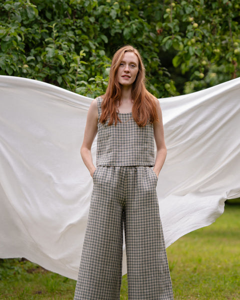 Model is posing so the front of the green gingham culottes is fully visible. Such a pose accentuates the side pockets as a feature. Behind her is a white sheet.