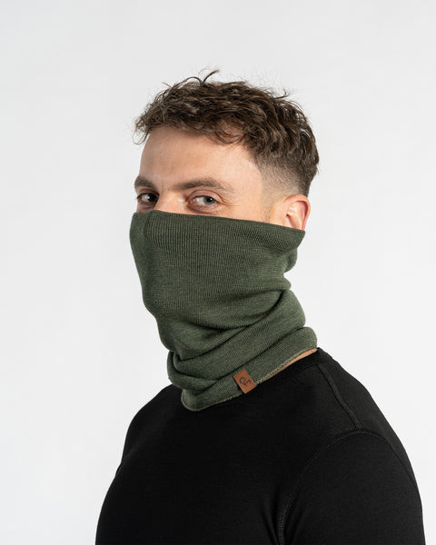 A man with short, dark hair and light eyes looks toward the camera while modeling a dark green knitted neck gaiter. The gaiter is pulled up over his nose and mouth, covering the lower portion of his face, and is set against a plain white background.